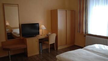 Ein Hotelzimmer mit einem Bett, einem Schreibtisch mit Fernseher, einem Sessel, einem Kleiderschrank und einem Fenster mit Vorhängen.
