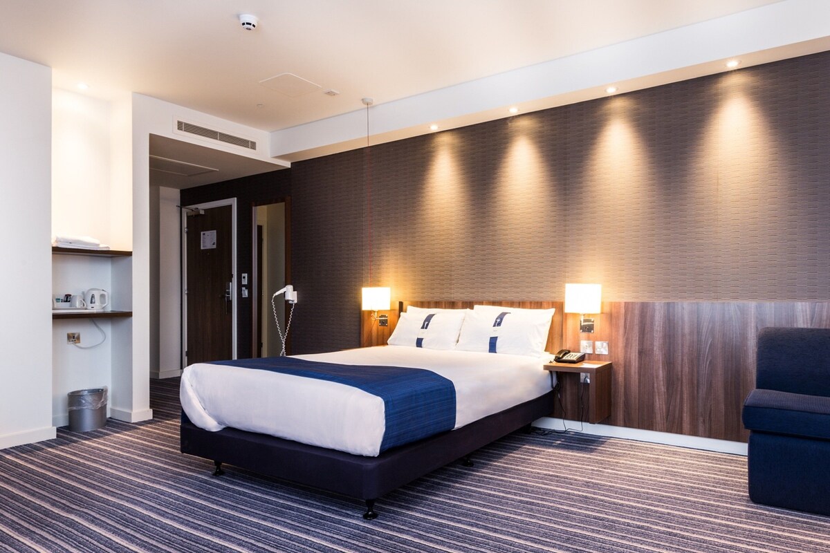 A hotel room with a large bed, bedside lamps, a chair, a desk, and a wall-mounted heater.