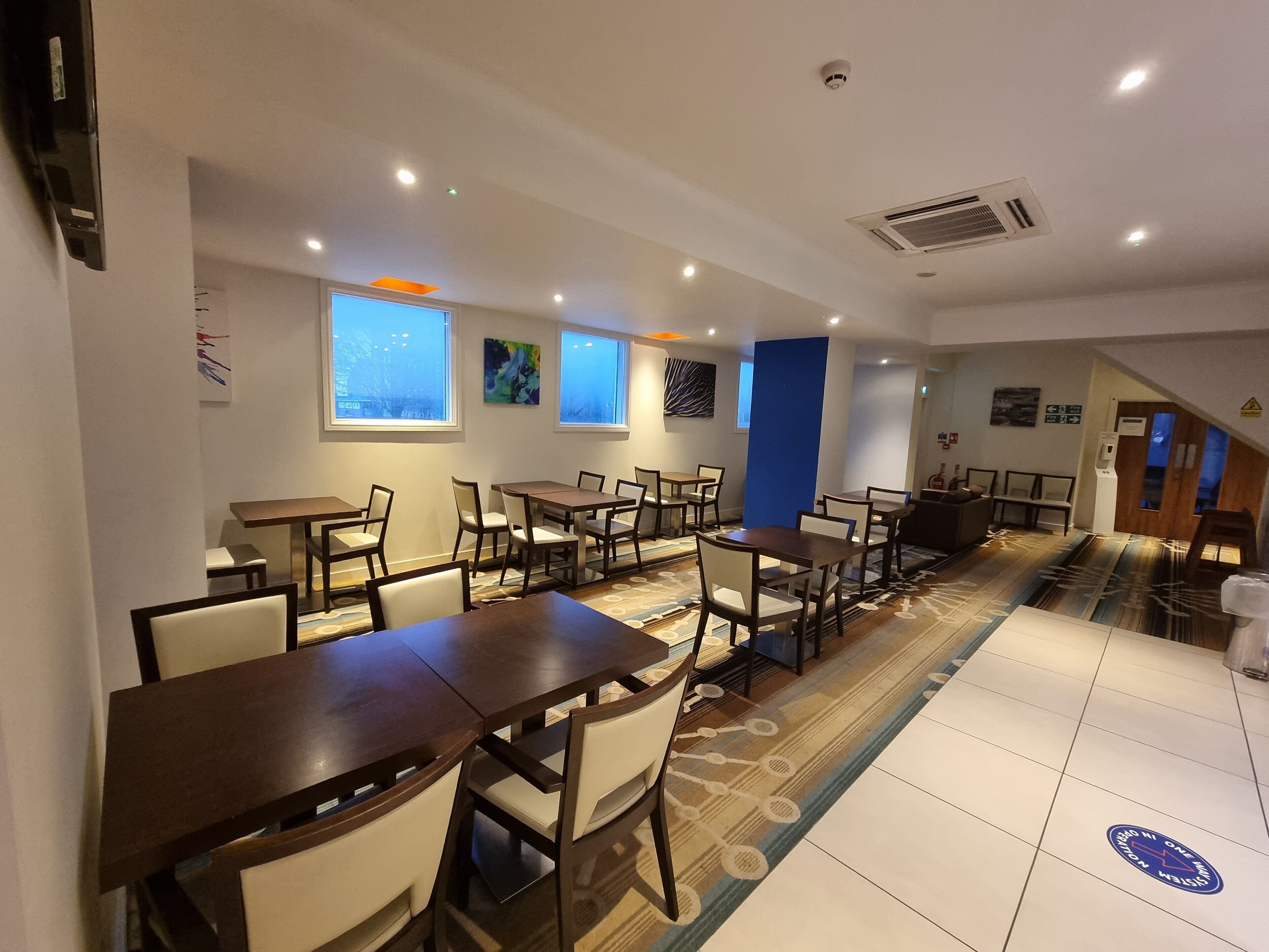 A hotel dining area with multiple tables and chairs, large windows, and a patterned carpet.