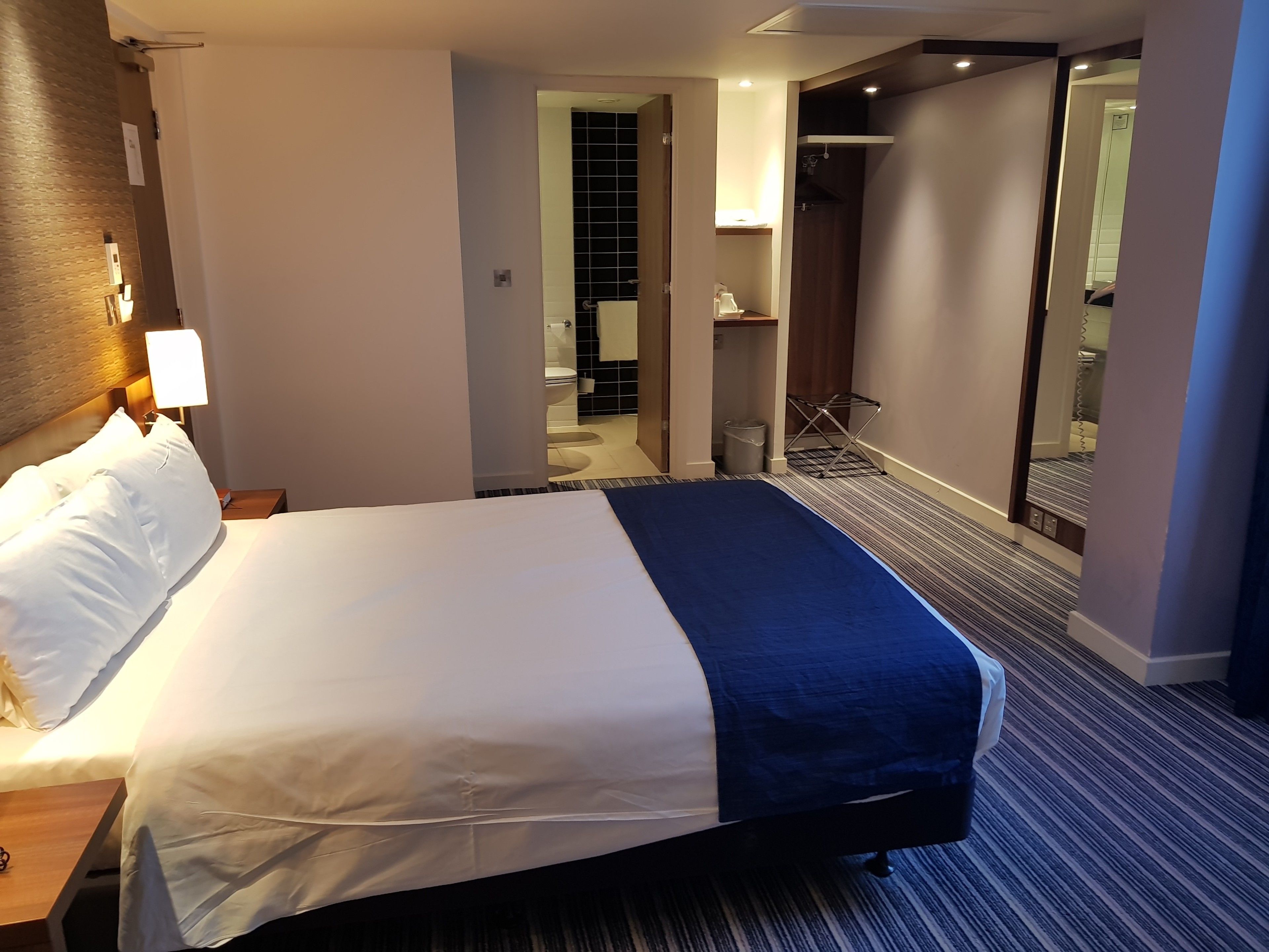 A hotel room with a bed, a nightstand, a lamp, a mirror, a bathroom visible through an open door, and a small seating area with a chair and a table.