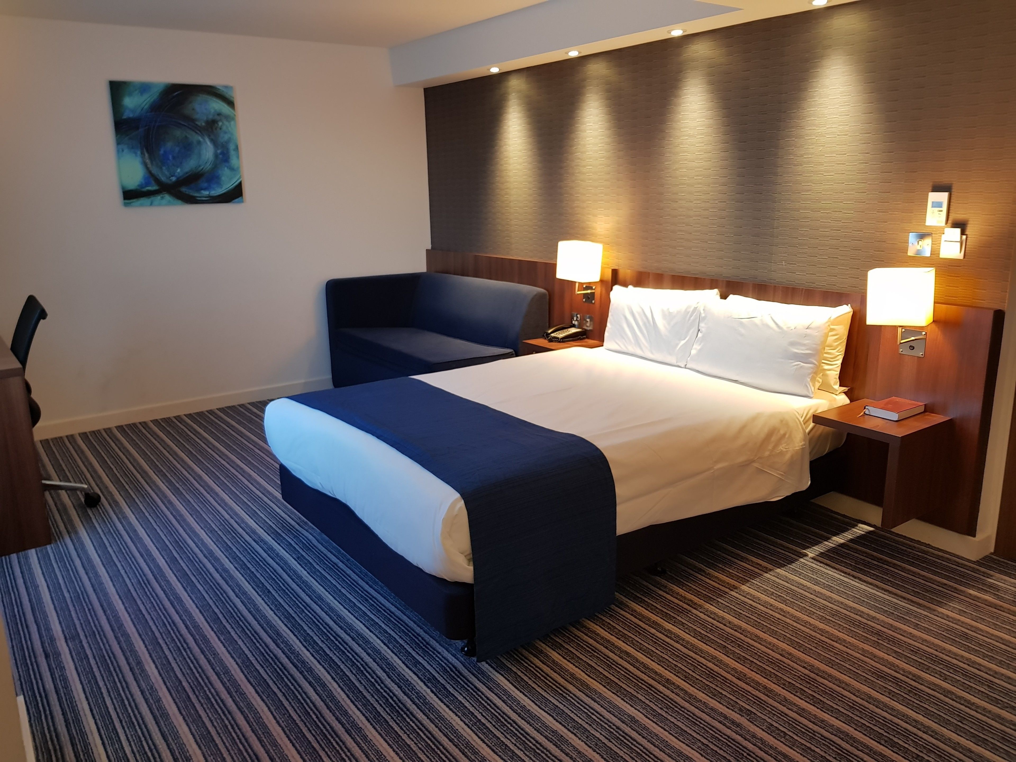 Una habitació d'hotel amb un llit gran, un sofà blau, una taula de nit amb una làmpada i un quadre a la paret.