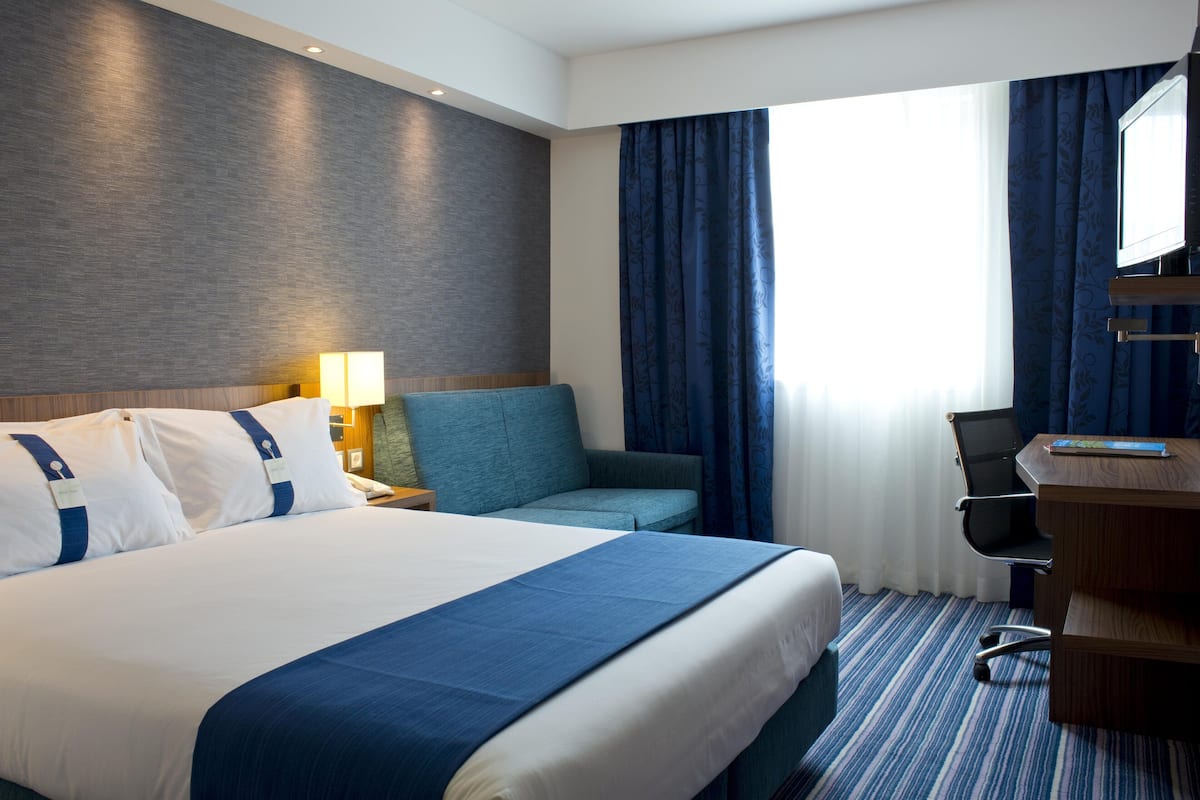 Habitación de hotel con una cama grande, un escritorio, una silla, una lámpara y una ventana con cortinas.