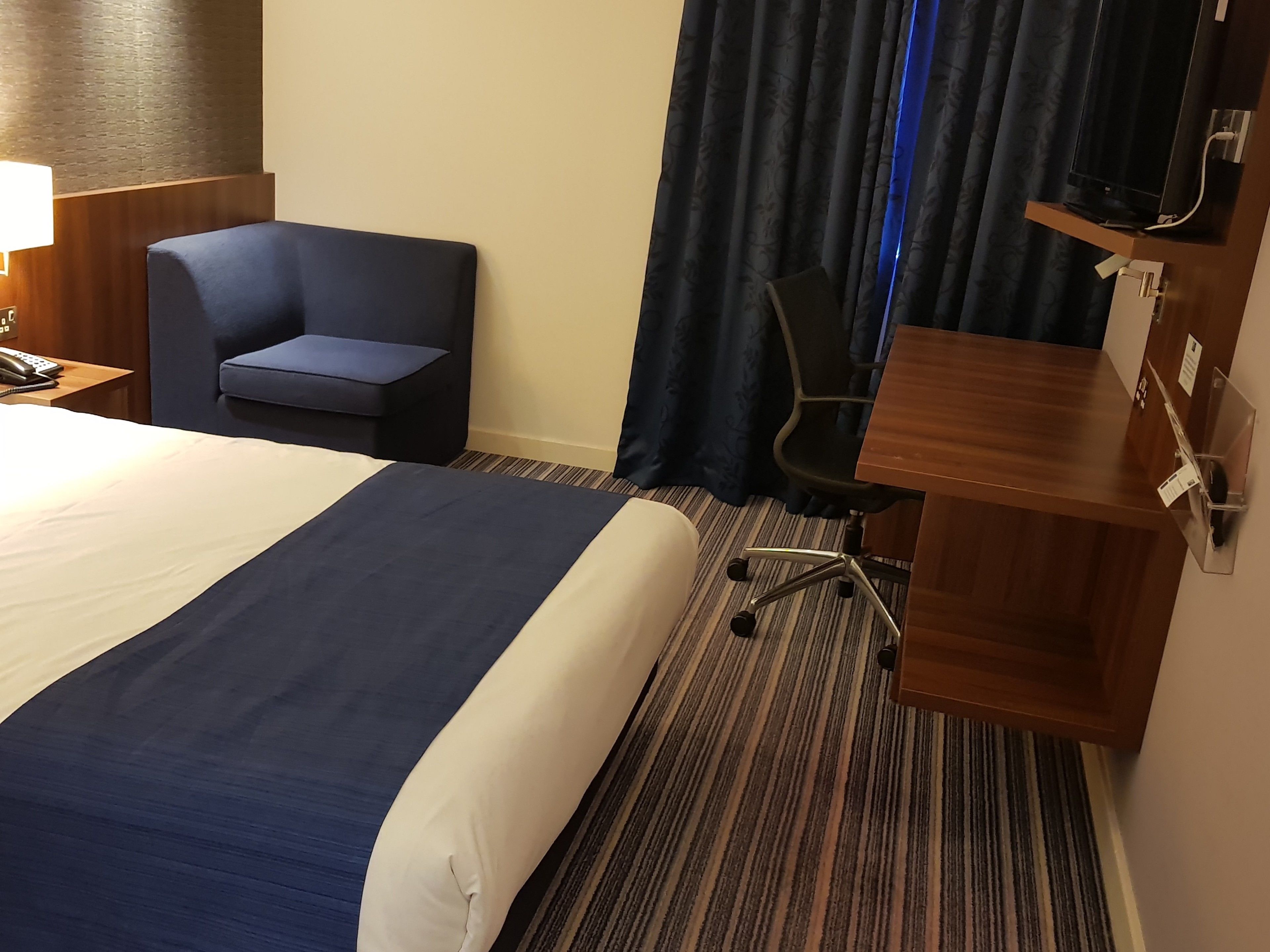 A hotel room with a large bed, a desk, a chair, a lamp, and a curtain.