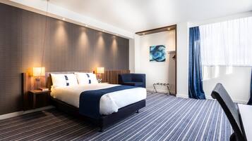 Una habitación de hotel con una cama, una silla, un escritorio, un cuadro y un ventanal con cortinas.
