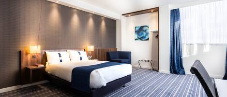 Ein Hotelzimmer mit einem Bett, einem Sessel, einem Schreibtisch, einem Gemälde und einem fenster mit Vorhängen.