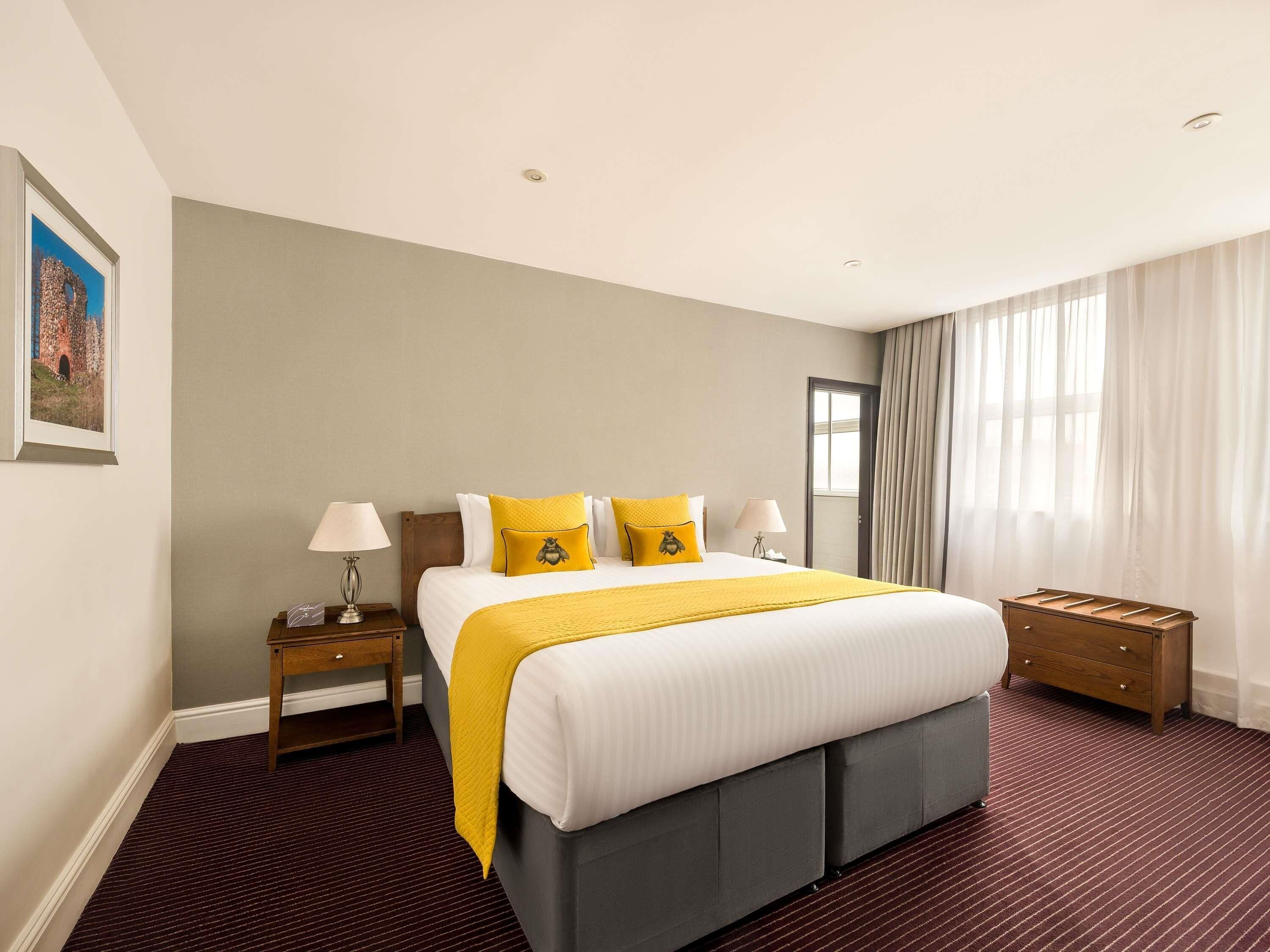 A hotel room with a large bed, two bedside tables, a wooden nightstand, a mirror, and a window with curtains.