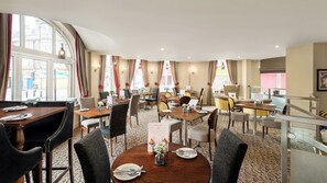 Un comedor de hotel con varias mesas y sillas, amplias ventanas con cortinas y suelo alfombrado.