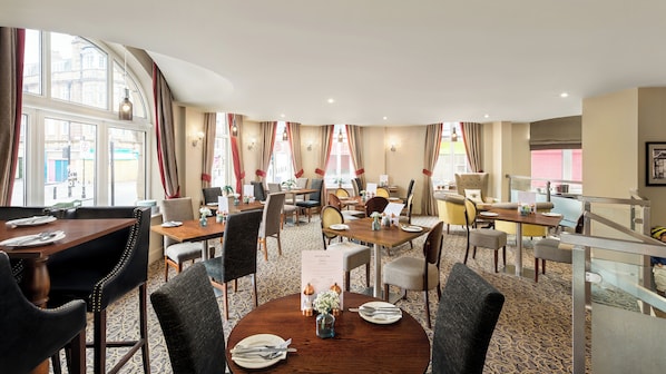 Un comedor de hotel con varias mesas y sillas, amplias ventanas con cortinas y suelo alfombrado.