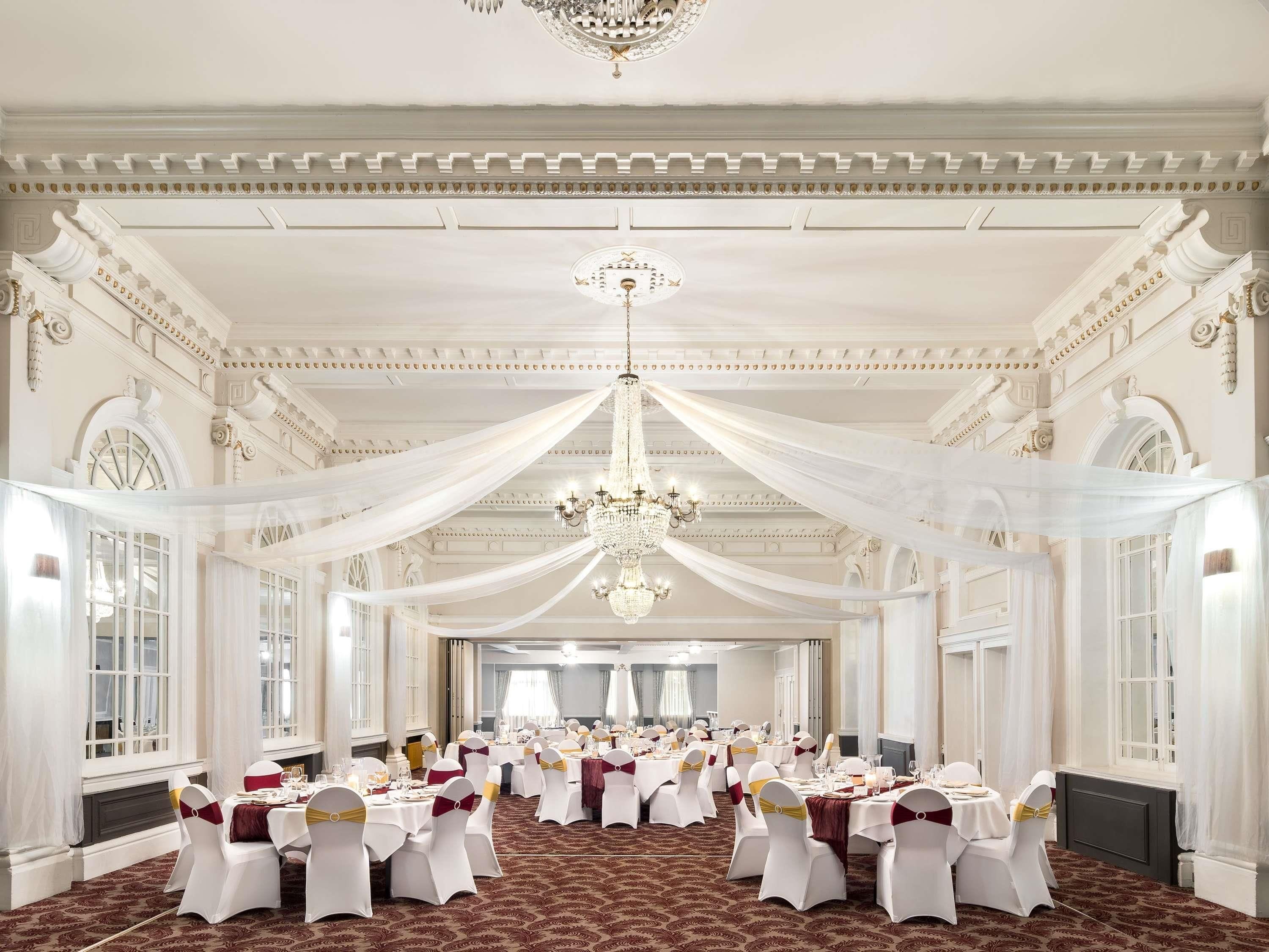 Un salón de banquetes con mesas dispuestas para un evento formal, cubiertas con tela blanca, y una araña colgando del techo.