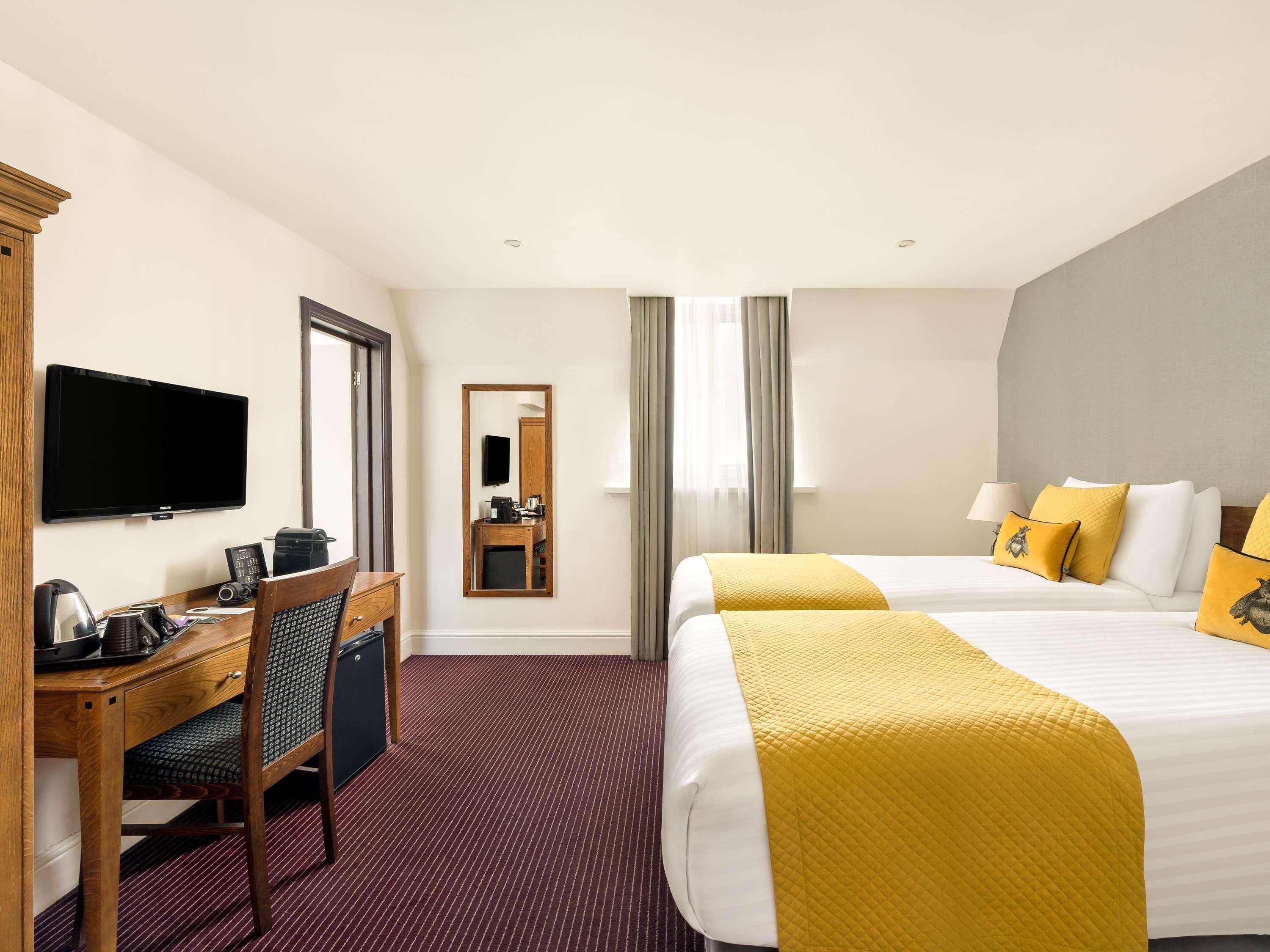 A hotel room with a desk, a TV, a bed with yellow bedding, and a mirror.