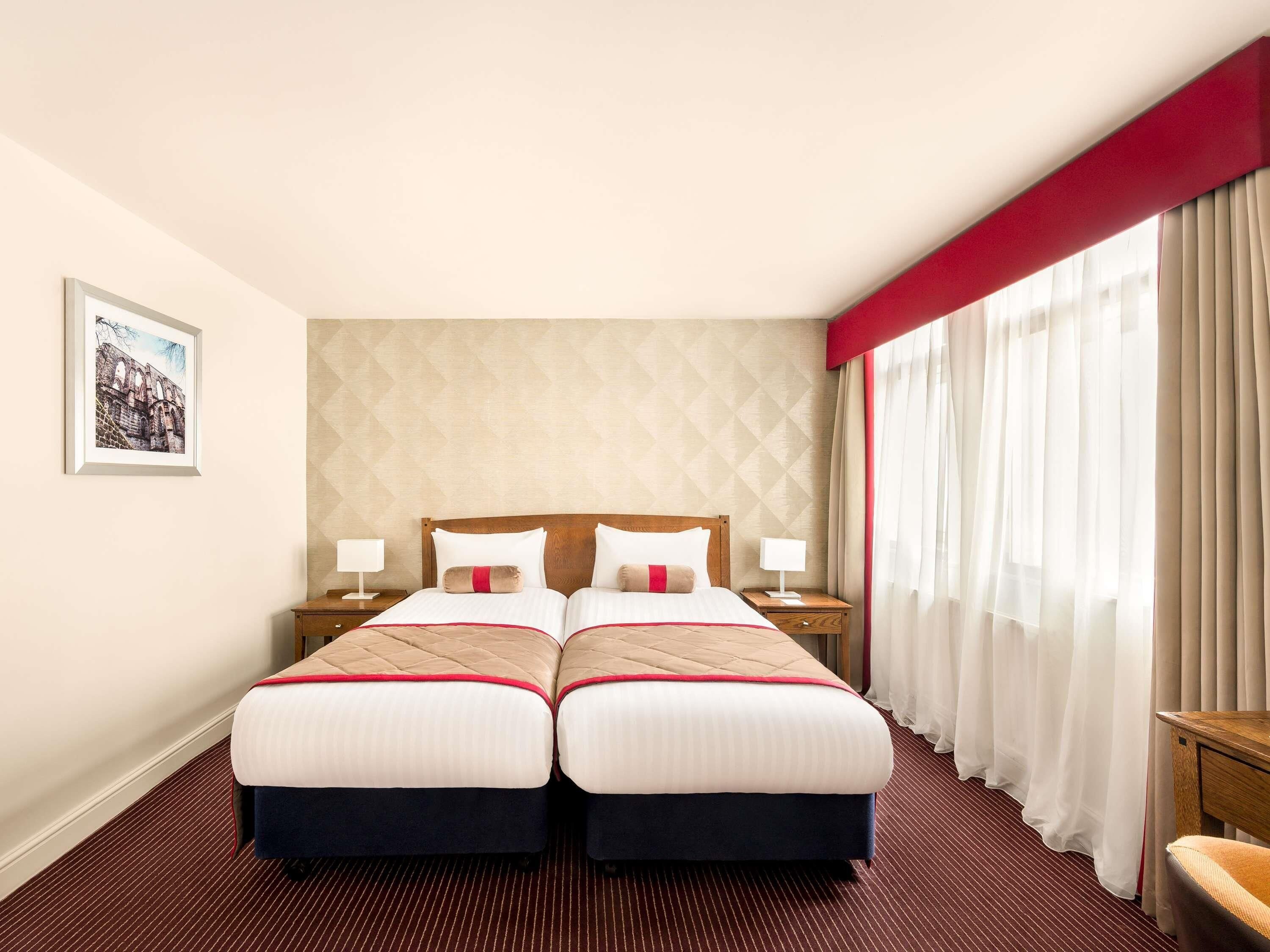 Habitación de hotel con dos camas, un ventanal con cortinas, un cuadro enmarcado en la pared y una pared de acento roja.