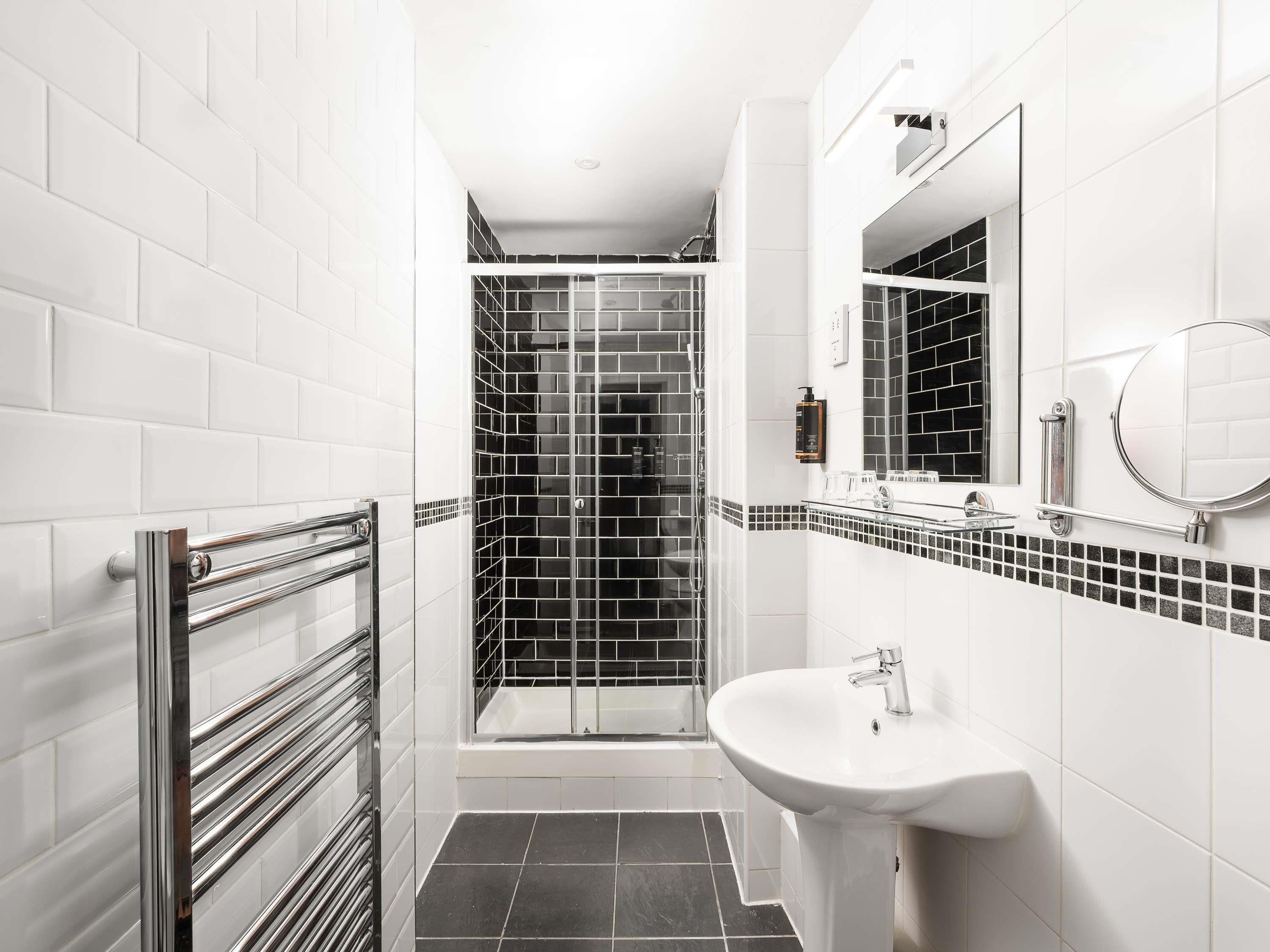 A modern bathroom with white subway tiles, a black and white mosaic shower, a glass shower enclosure, a white sink with a chrome faucet, and a large mirror.