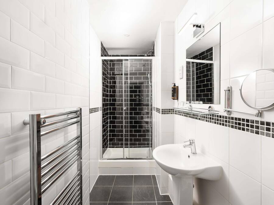 Un bagno moderno con piastrelle bianche tipo metro, una doccia in mosaico bianco e nero, box doccia in vetro, lavandino bianco con rubinetto cromato e uno specchio grande.