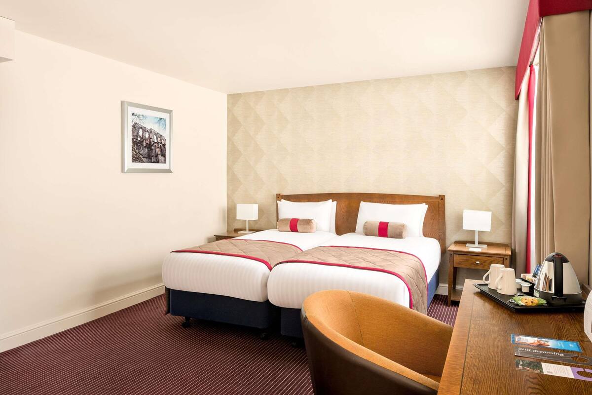 Een hotelkamer met een tweepersoonsbed, twee nachtkastjes, een stoel, een bureau met dienblad, en een ingelijst schilderij aan de muur.