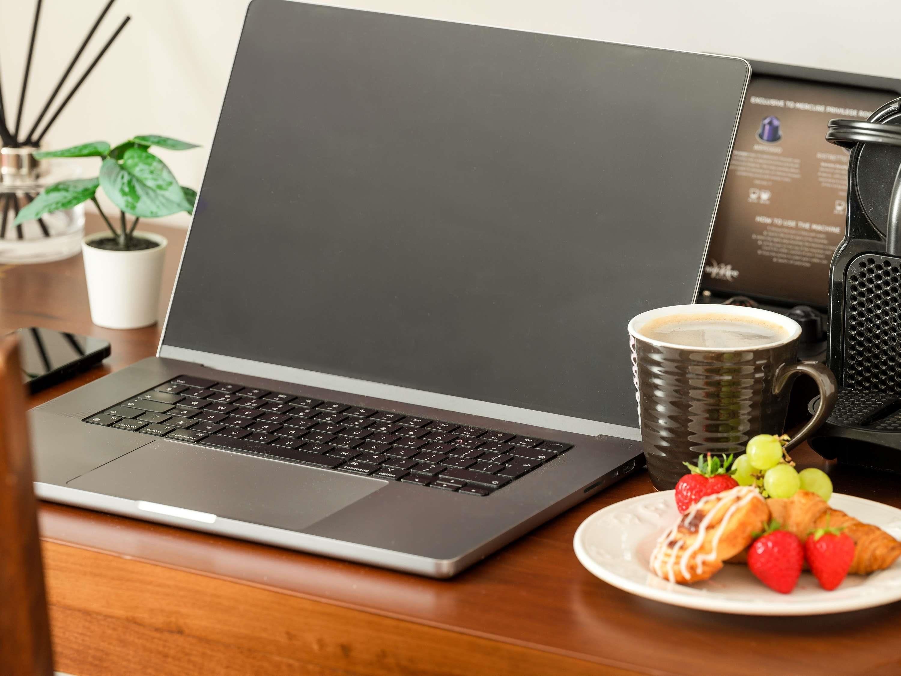 Una laptop sobre un escritorio de madera con una taza de café, un plato de comida y un teléfono móvil.