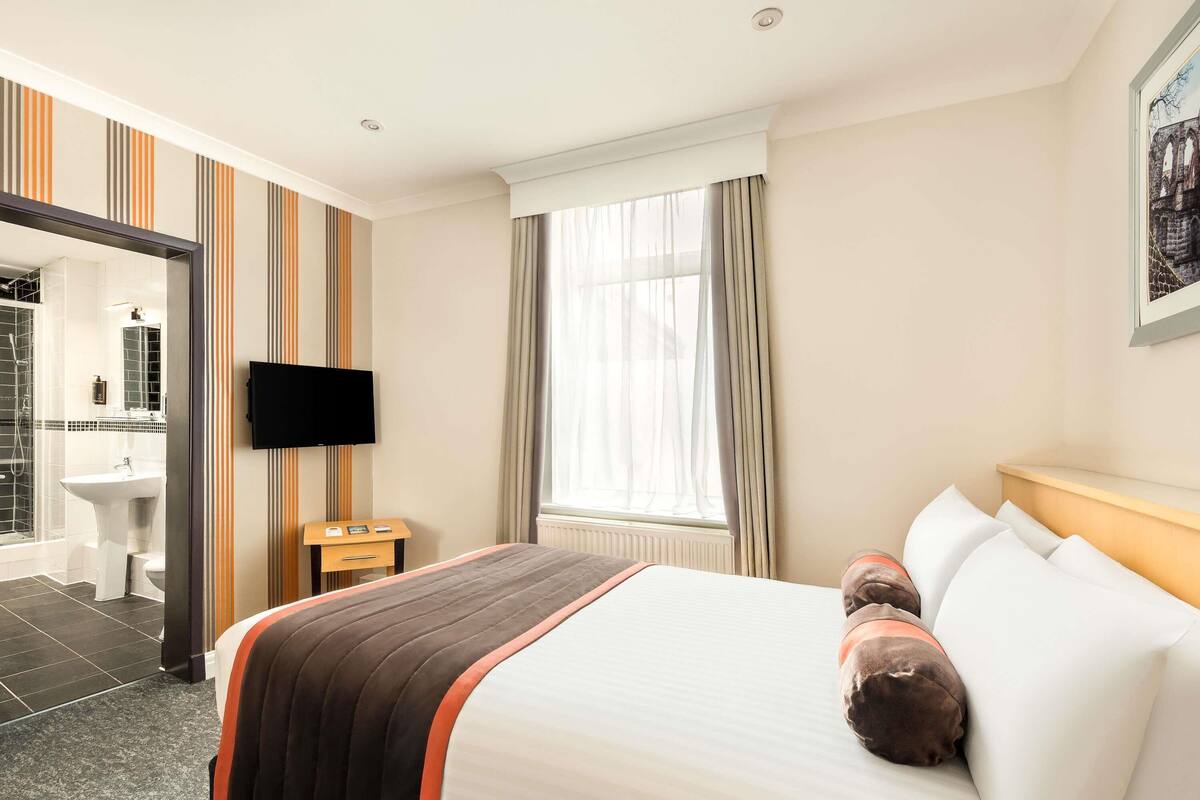 Hotelkamer met een bed, televisie, raam met gordijnen, badkamer met douche en wastafel, en een schilderij aan de muur.