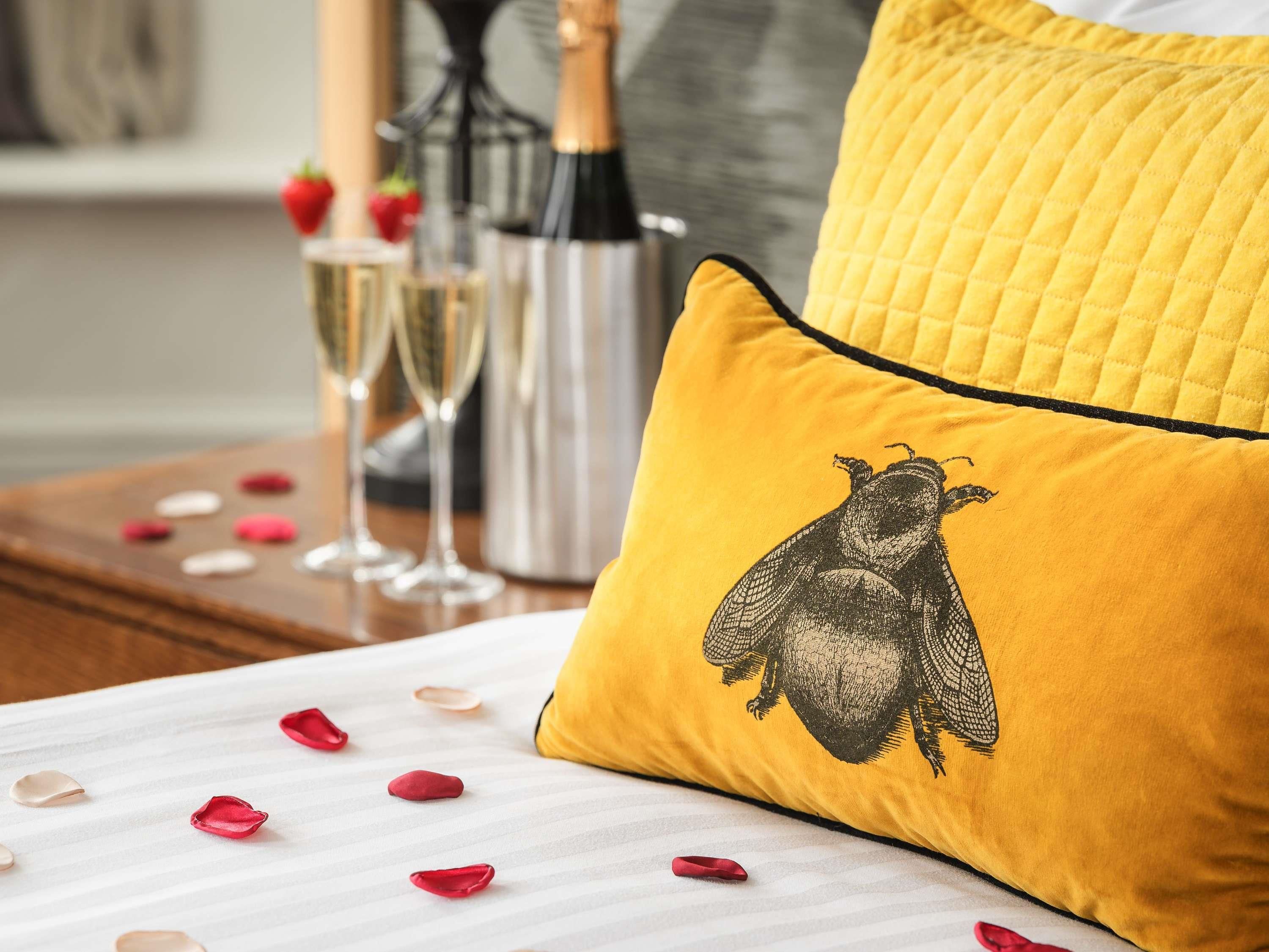 Una cama con un almoháque amarillo que muestra un abejorro bordado en negro, una colcha blanca y pétalos de rosa dispersos.