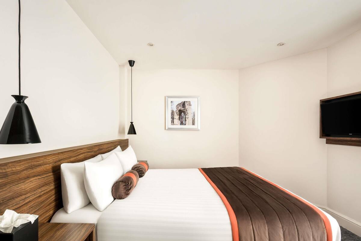 Een hotelkamer met een bed, nachtkastje, lamp, een foto aan de muur en een aan de wand bevestigde televisie.