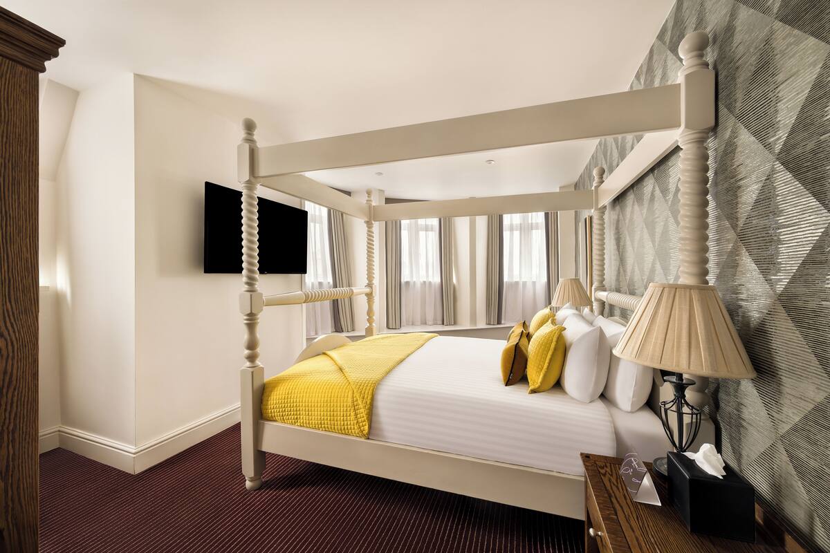 Een bed met baldakijn, een wit hoofdeinde, een flatscreen televisie aan de muur en behang met een patroon op de aangrenzende muur.