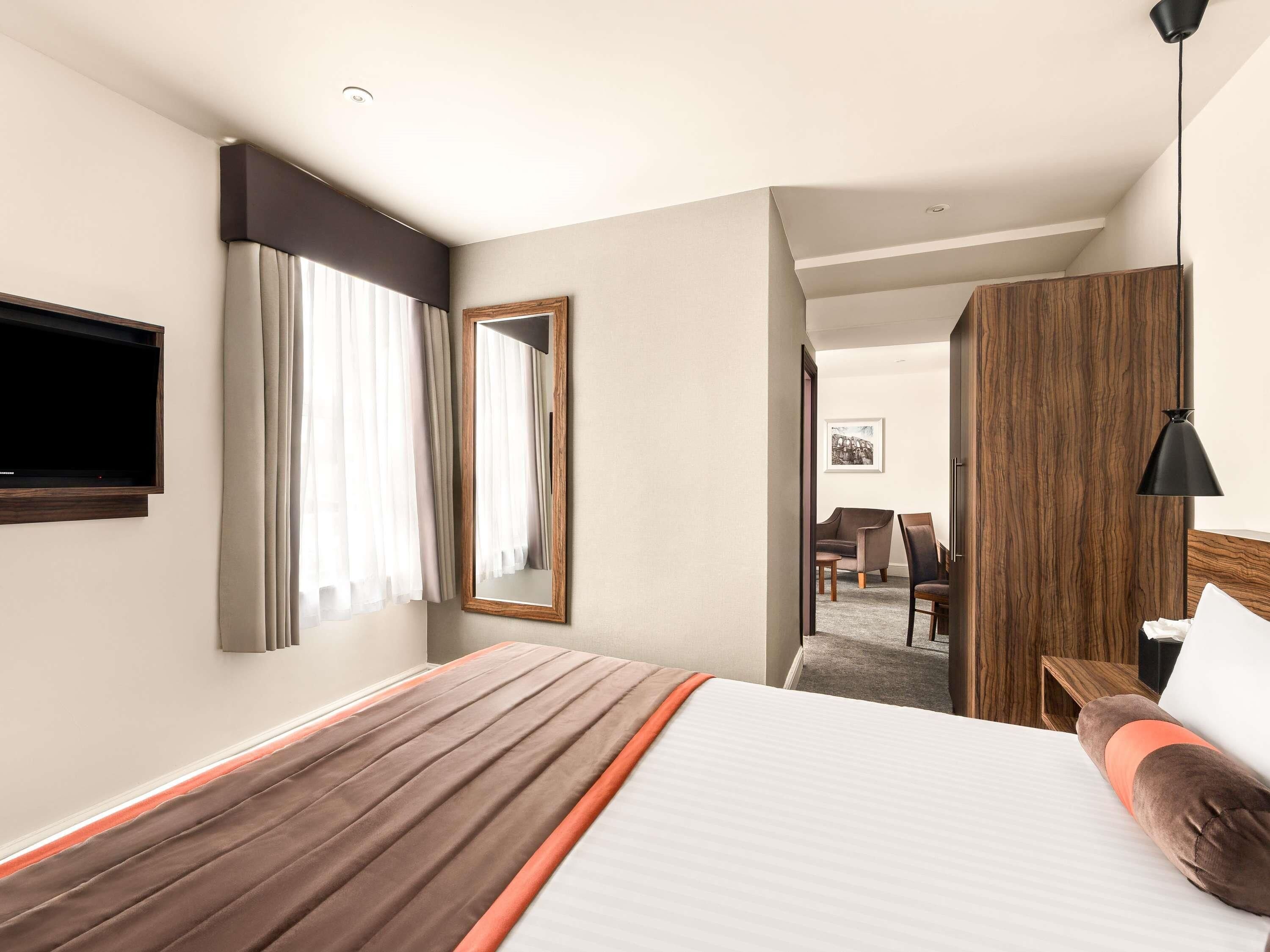 A hotel room with a large bed, a flat-screen TV, a wooden headboard, a mirror, and a desk with a chair.