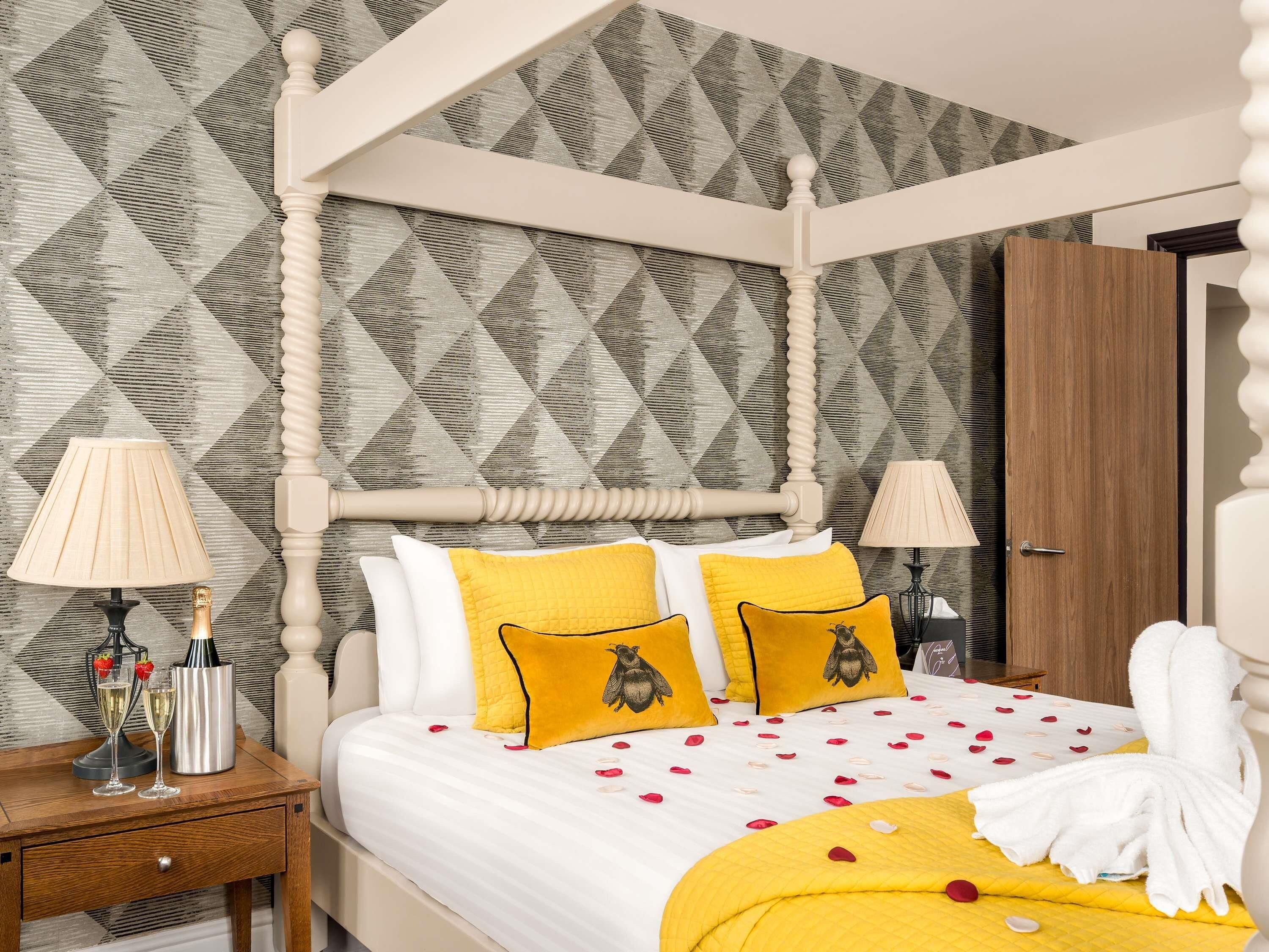 A four-poster bed with a geometric patterned wallpaper, two bedside lamps, and a wooden nightstand with a bottle and glasses.