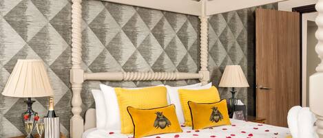 A four-poster bed with a geometric patterned wallpaper, two bedside lamps, and a wooden nightstand with a bottle and glasses.
