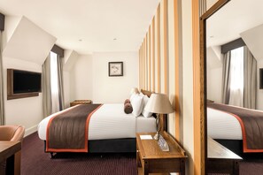Una habitación de hotel con una cama grande, una mesita de noche de madera, un escritorio y un espejo.