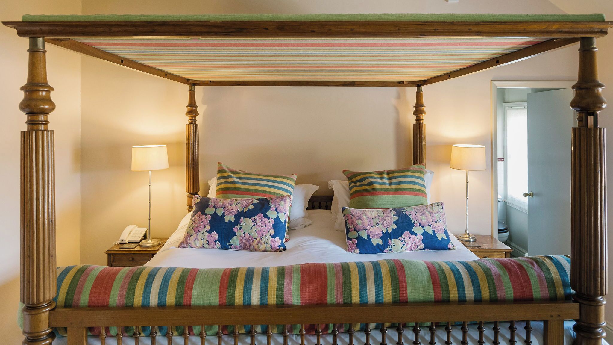 A four-poster bed with floral pillows, striped bedspread, and bedside lamps.