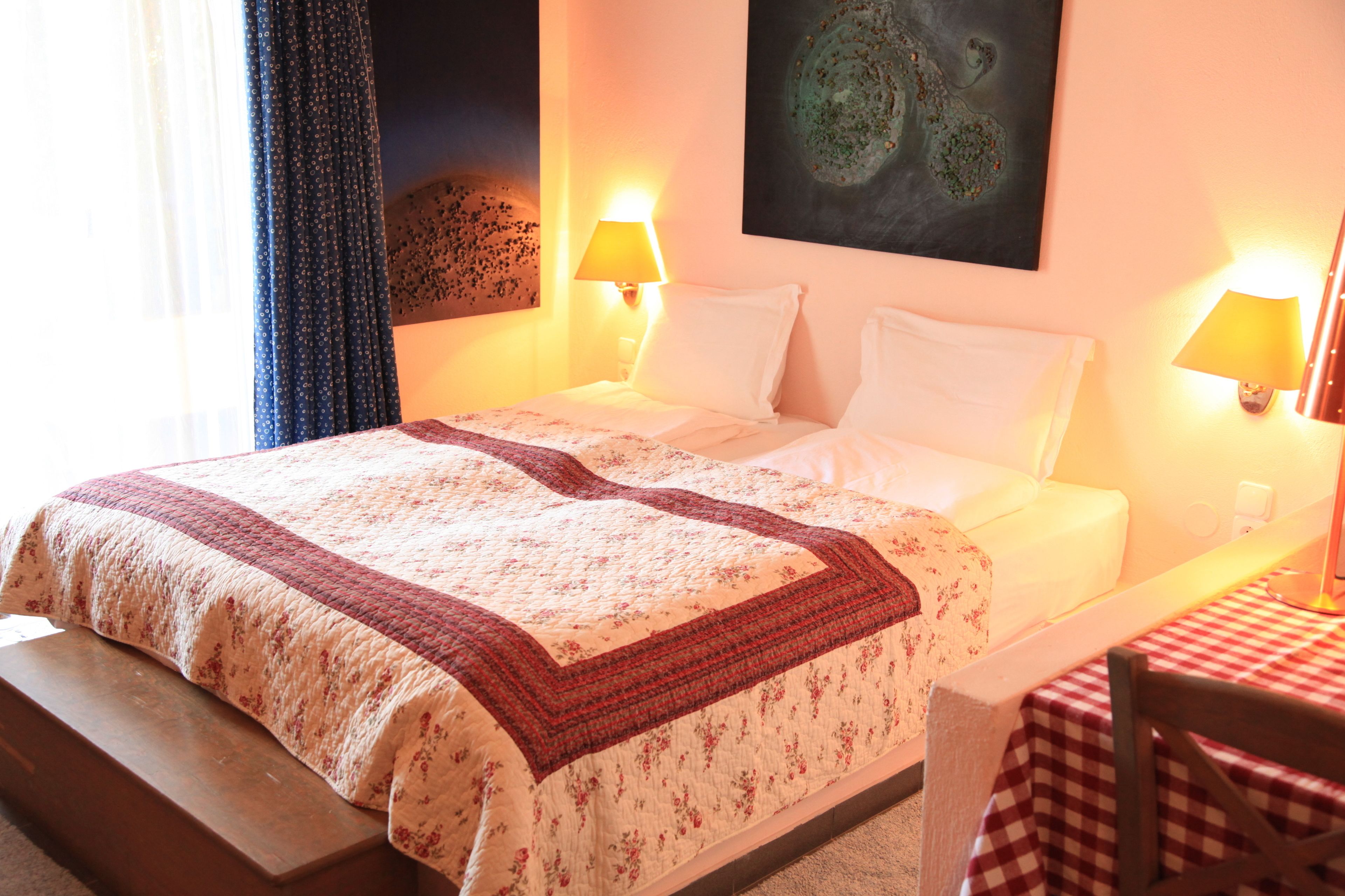 Une chambre d’hôtel avec un lit, deux lampes de chevet, un tableau au mur et une nappe à carreaux sur une petite table.