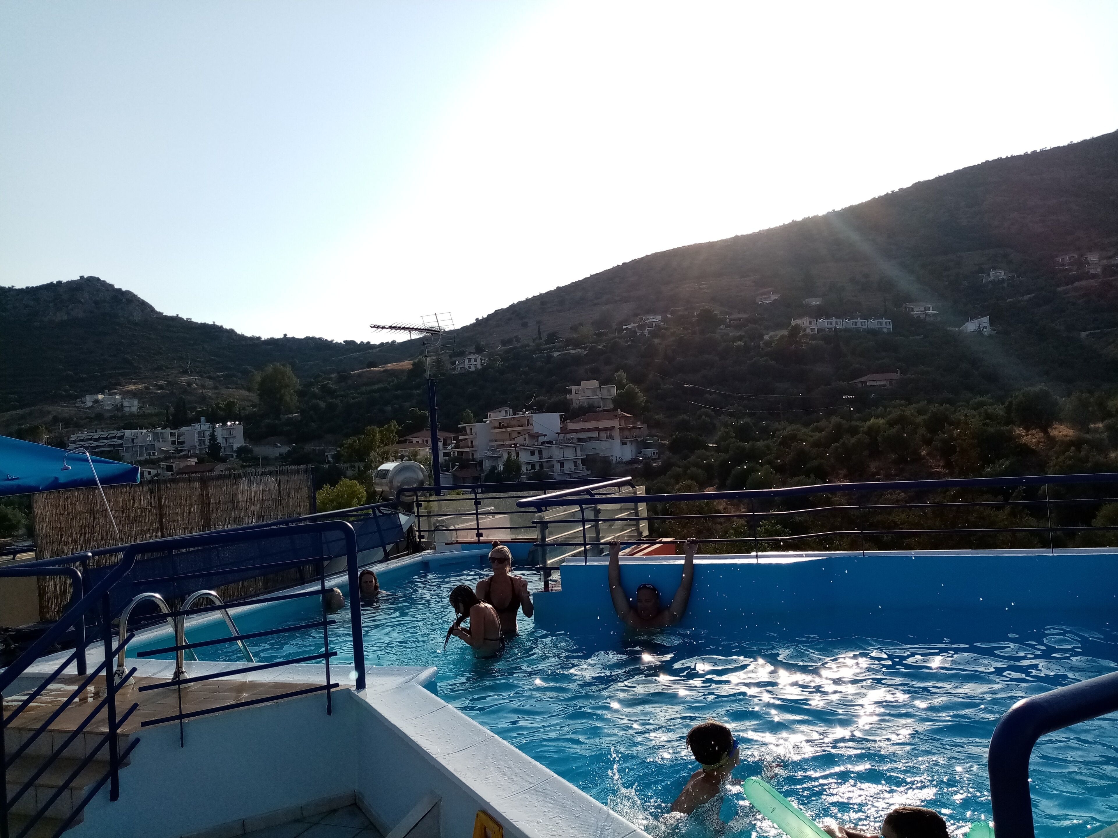 A swimming pool with people enjoying the water, surrounded by a hilly landscape and buildings in the background.