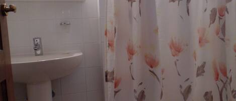 Un baño con un lavamanos blanco, un espejo encima y un inodoro. Se ve parcialmente una cortina de ducha con estampado floral.