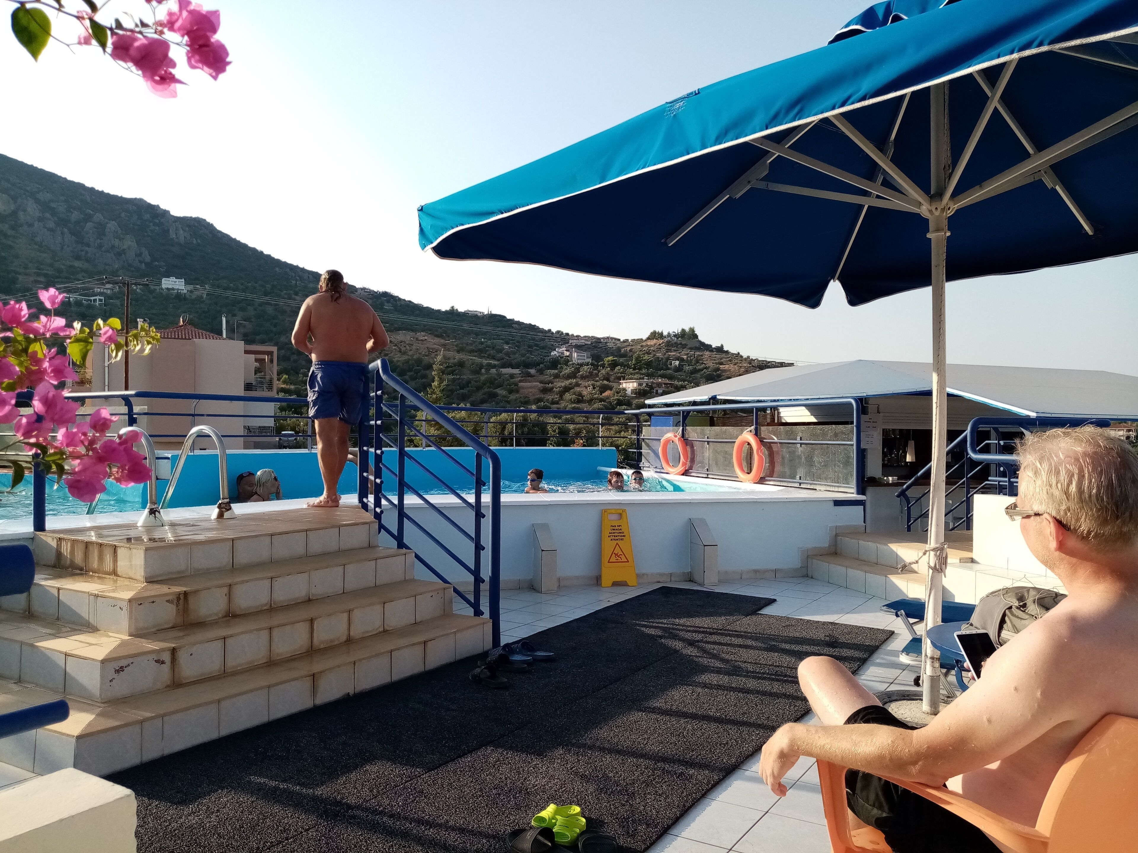 A pool area with steps leading into the water, a blue umbrella, and a person sitting on a chair.