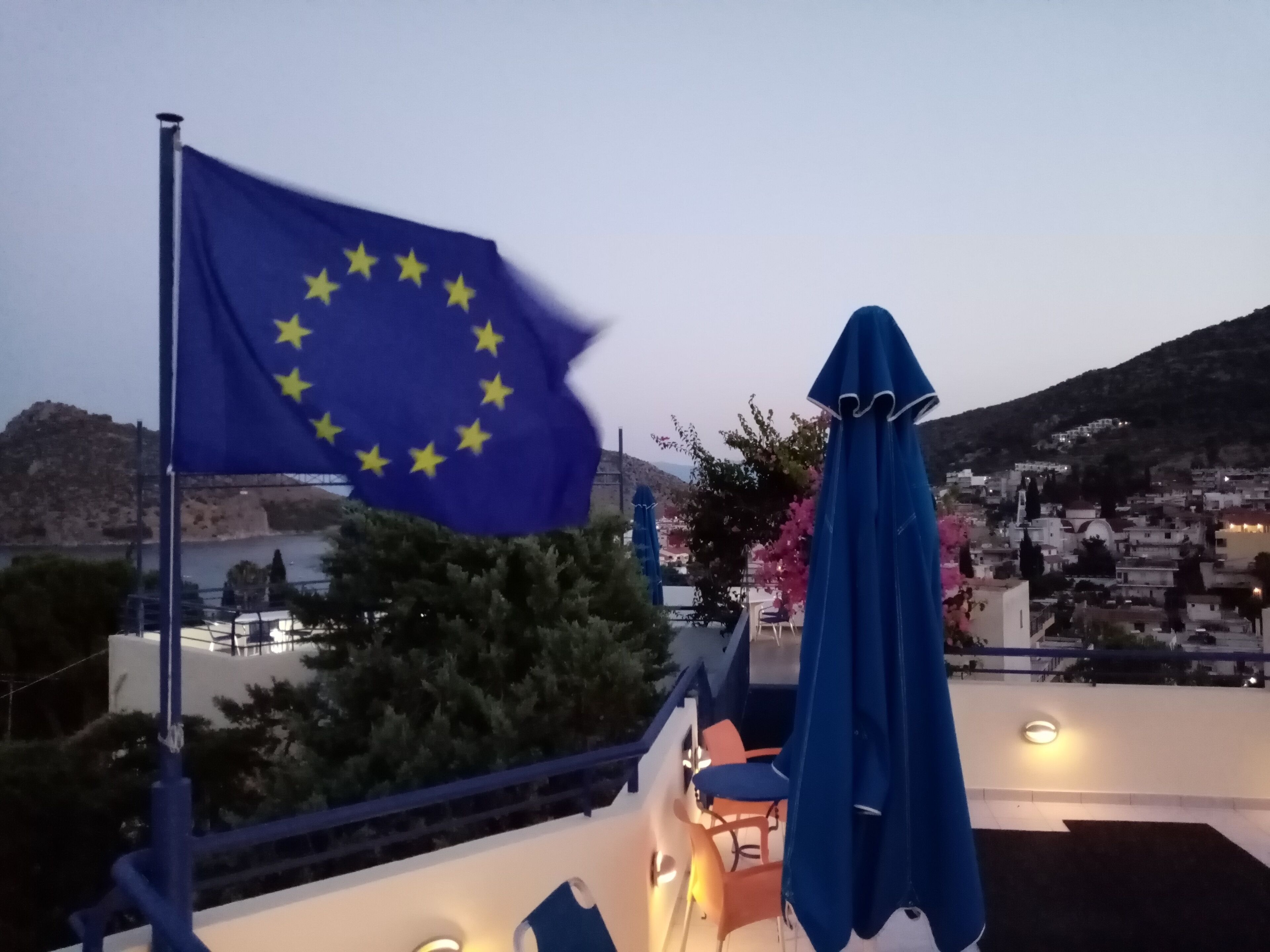 A flagpole with the European Union flag, outdoor seating with umbrellas, and a view of a coastal town with buildings and hills in the background.