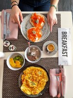 Desayuno con salmón ahumado sobre tostada, un cuenco de frutas variadas, un bol de semillas y frutos secos, una taza de café y una tortilla en un plato.