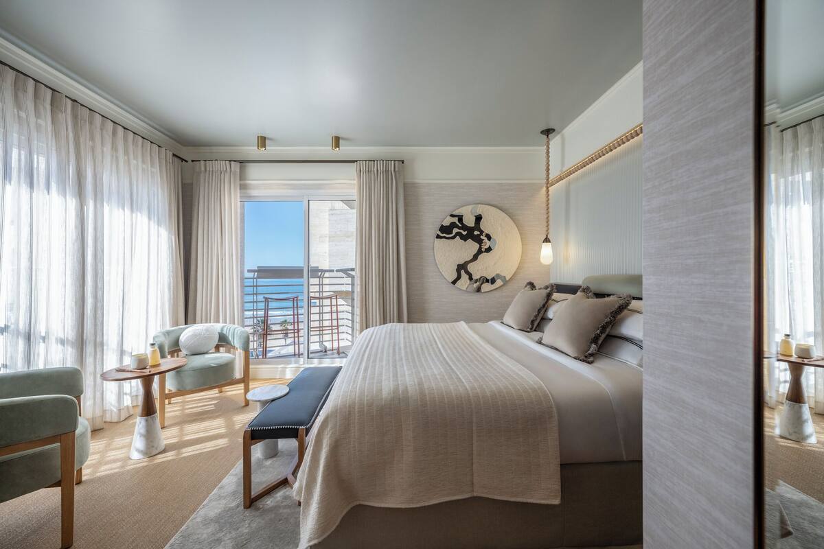 A modern hotel room with a large bed, a seating area with a bench and chairs, a view of the sea, and a decorative wall piece.