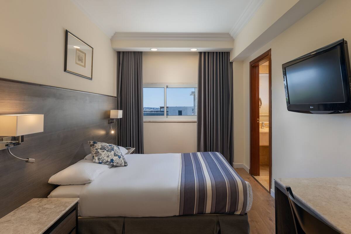 Ein Hotelzimmer mit Bett, Nachttischlampen, einem Flachbildfernseher, einem Fenster mit Vorhängen und einer Tür zu einem Badezimmer.