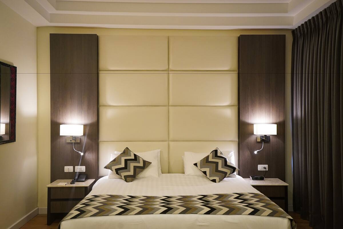 Ein Hotelzimmer mit einem großen Bett, zwei Nachttischen mit Lampen, einer Kopfteil mit einer Lichtleiste und einem dunklen Vorhang am Fenster.