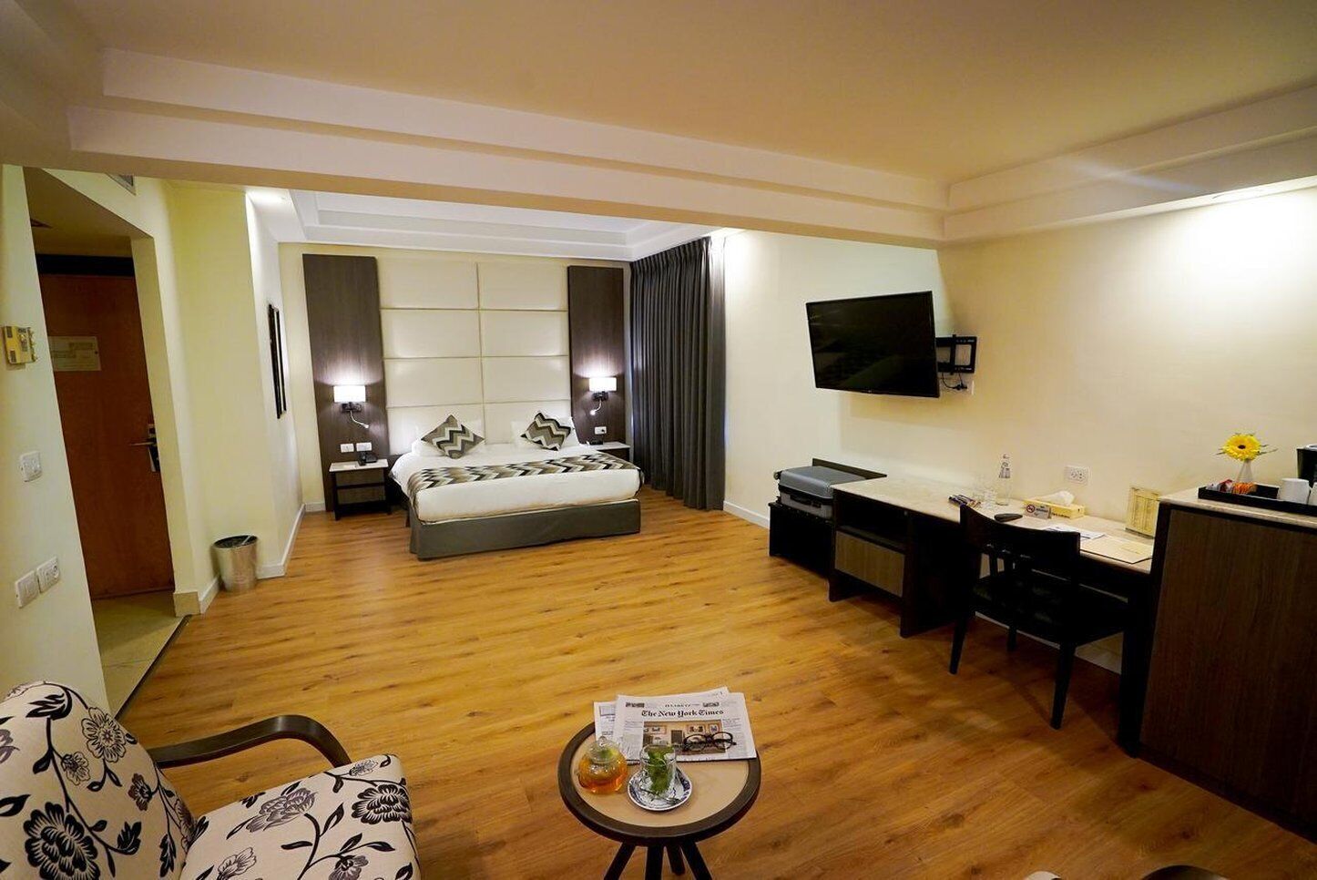 Habitación de hotel con cama, escritorio, silla, televisor y una mesita con un periódico y un vaso de agua.