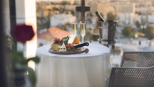 A table with a white tablecloth, a bottle of champagne, two glasses of champagne, a cheese board with various cheeses and fruits, a knife, and a cross. - Ambassador Hotel (Jerusalem)