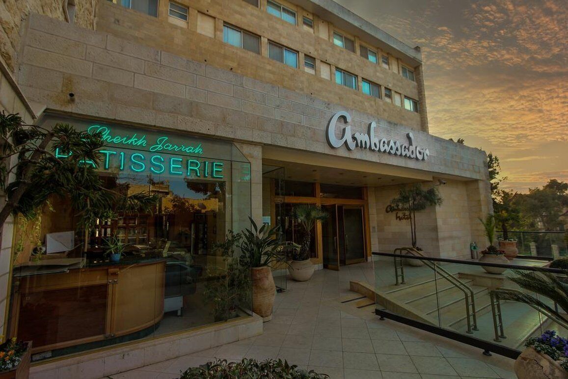 Un hotel con una fachada moderna, un restaurante visible en la planta baja llamado "Leikhn Jarruh TISSERIE", y un cartel con la leyenda "Ambassador Hotel" sobre la entrada.