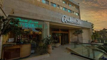 Un hotel con una fachada moderna, un restaurante visible en la planta baja llamado "Leikhn Jarruh TISSERIE", y un cartel con la leyenda "Ambassador Hotel" sobre la entrada.