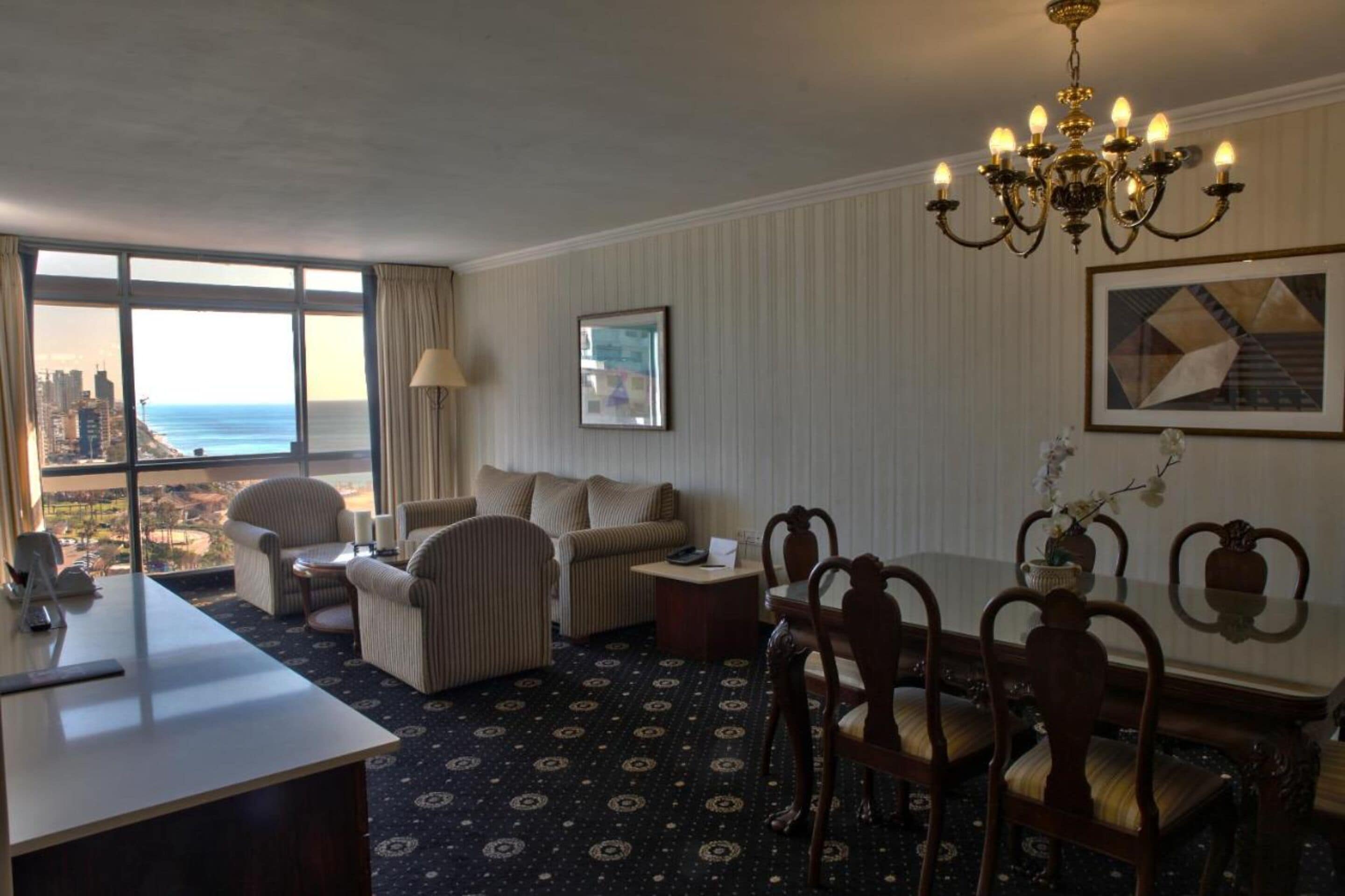 Quarto de hotel com uma grande janela com vista para a cidade e o oceano, área de refeições com um candelabro e carpete estampado.