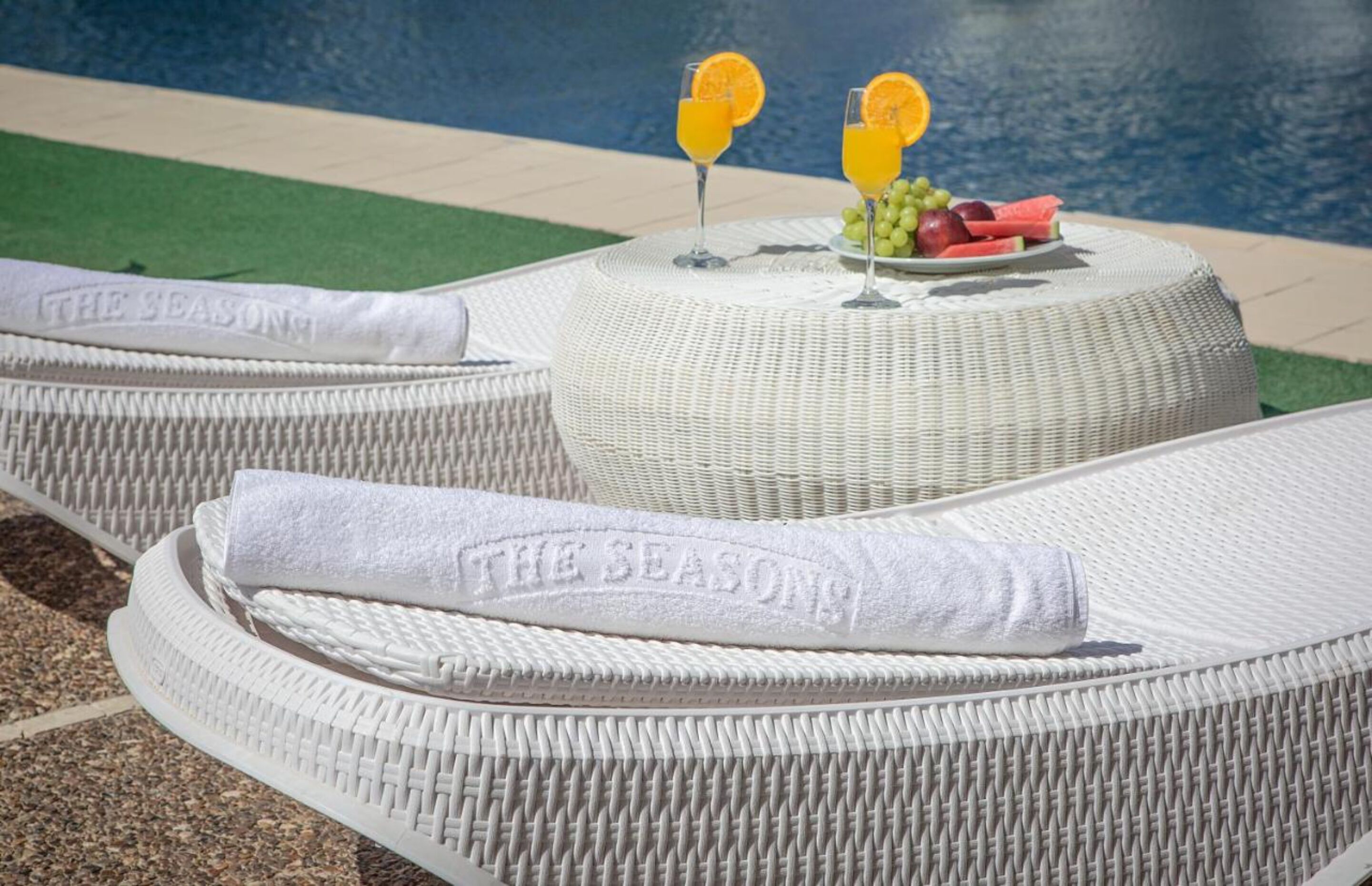 A poolside lounge area with wicker loungers and a round table with fruit and drinks.