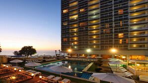 Un hotel con zona de piscina, tumbonas, sombrillas y vista al mar.