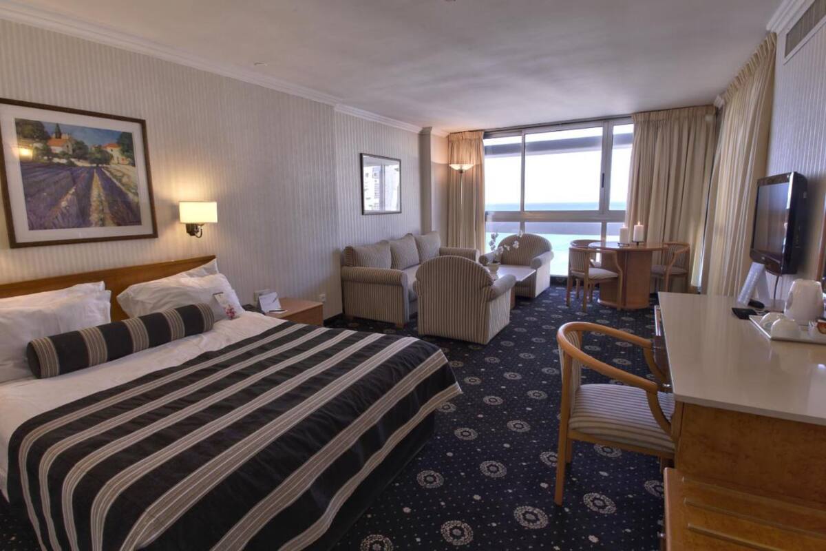 A hotel room with a large bed, a desk with a TV, a chair, and a view of the sea.