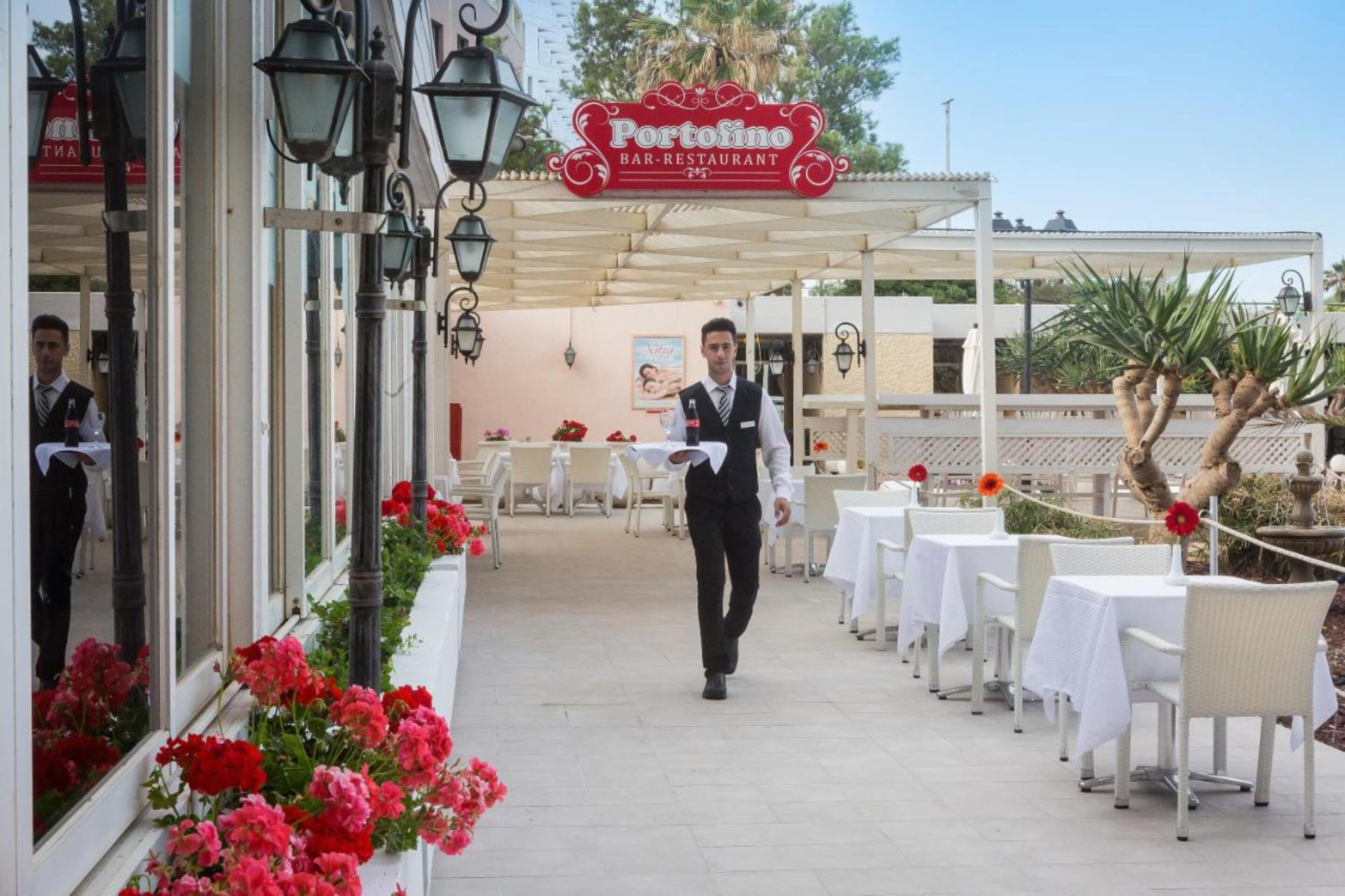 An outdoor restaurant with white tables and chairs, a waiter walking, and a sign that reads "Portofino Bar-Restaurant".