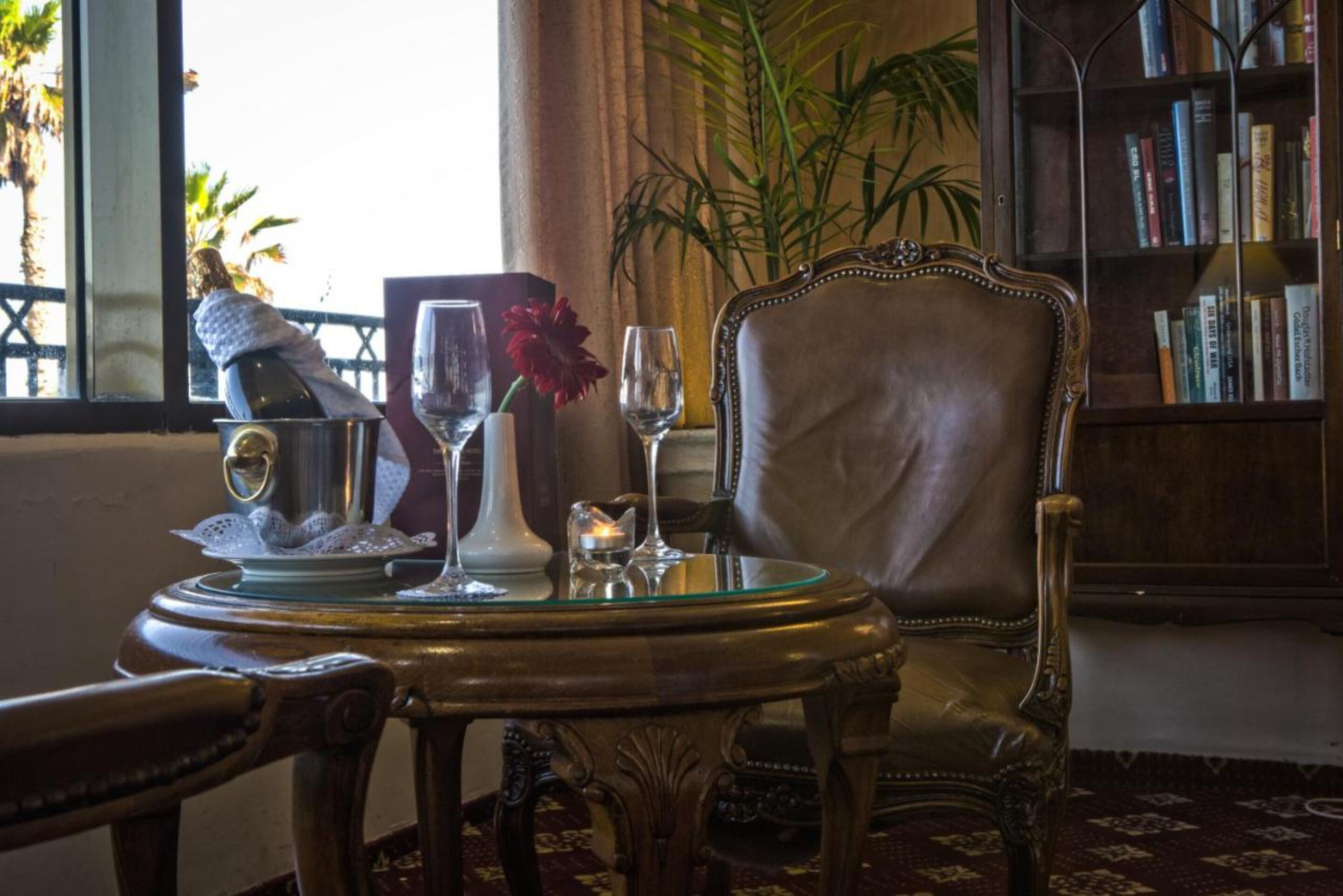 Una habitación con una mesa redonda dispuesta con una botella, vasos y una flor. Se ve una silla y una estantería al fondo.