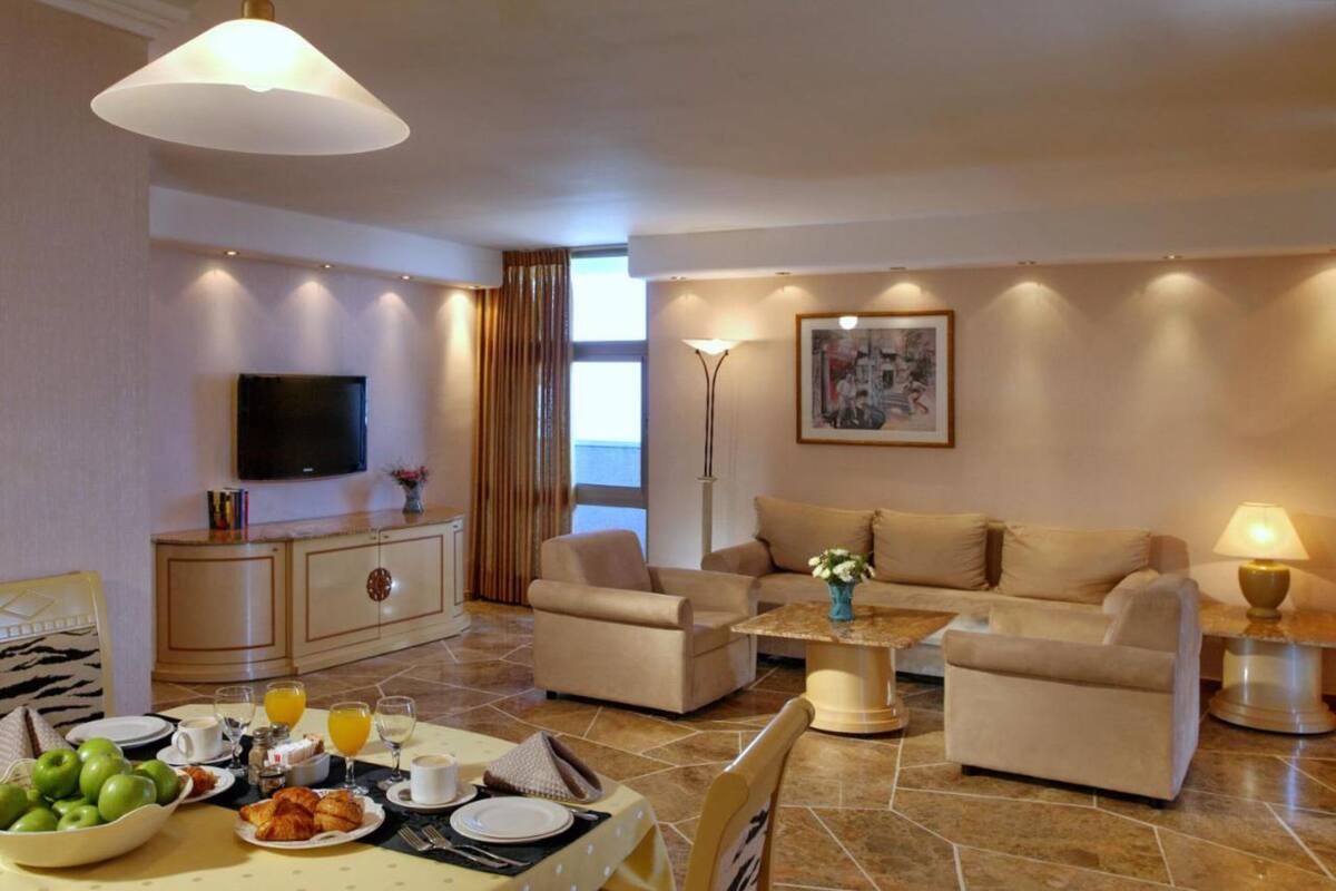 A modern living room with a sofa set, a dining table with breakfast items, a flat-screen TV, and a painting on the wall.