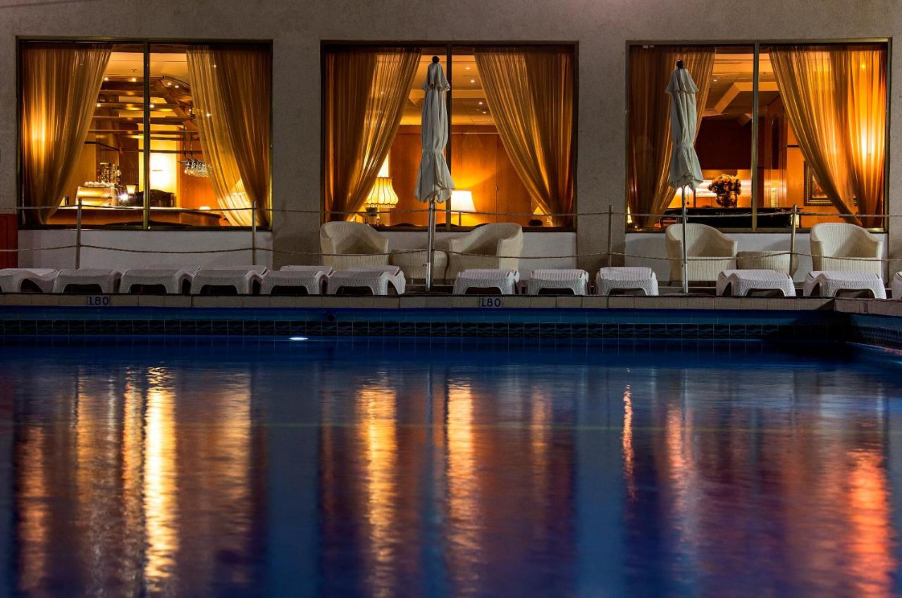 Zona junto a la piscina con sillones de descanso y sombrillas, una piscina y ventanas amplias con cortinas.