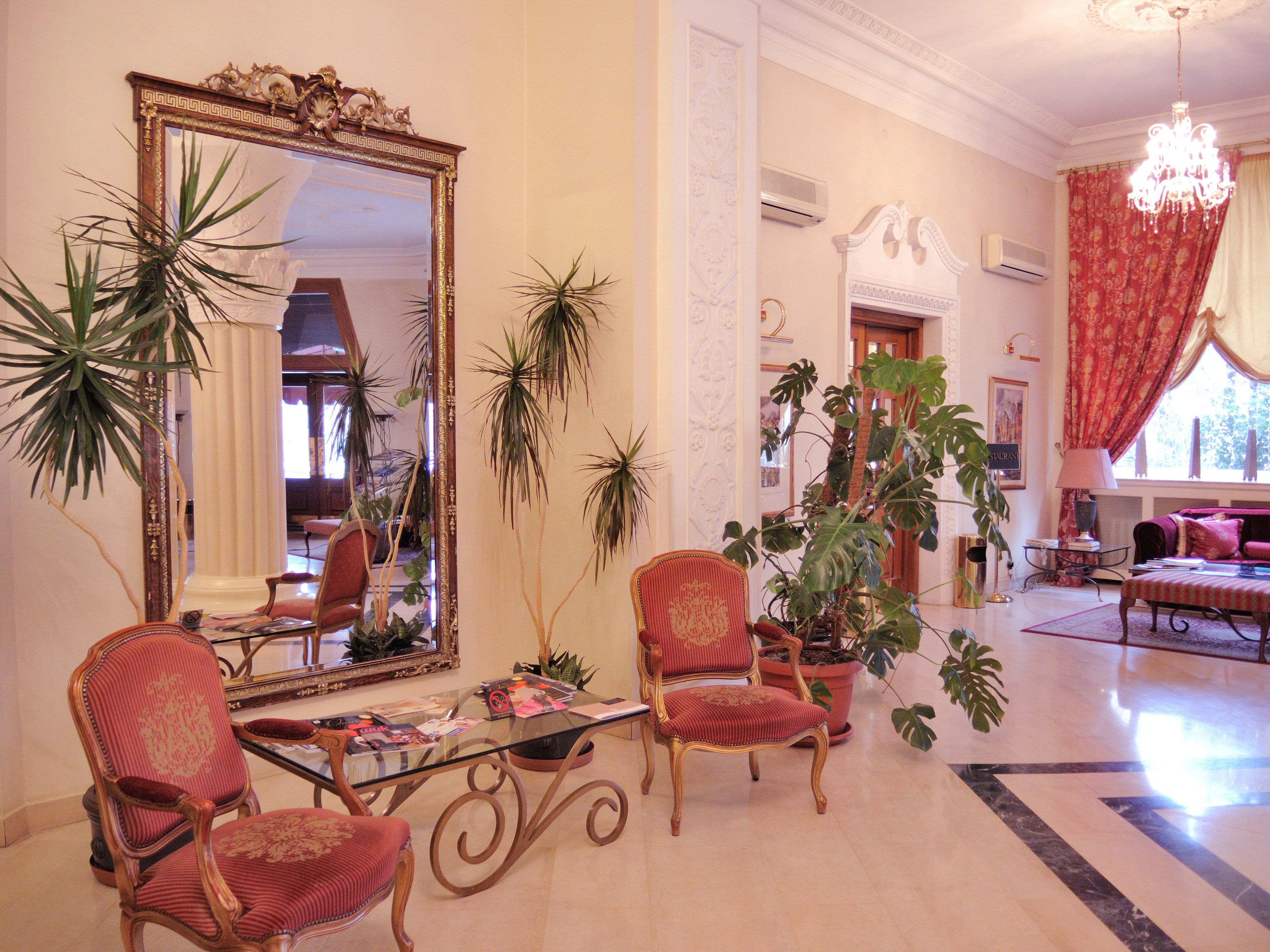 Un salón amplio con un espejo grande, dos sillones tapizados de rojo, una mesa de centro de vidrio y varias plantas en macetas.